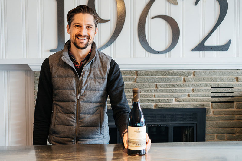 Brandon Thomas smiling and posing with a bottle of wine.