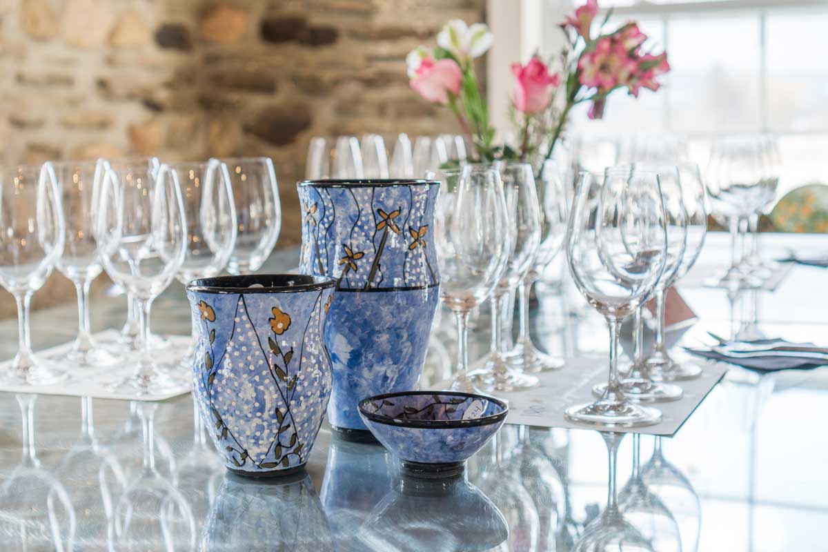 A table with three blue vases, several wine glasses, and flowers.